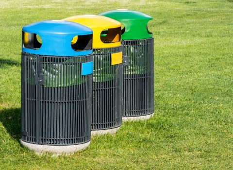Three cylindrical outdoor rubbish bins are placed on a well-maintained grassy area, each with a black plastic body featuring vertical slats and a rounded top with a distinct colour for the lid—blue on the left, yellow in the middle, and green on the right. The lids include oval-shaped openings for waste disposal, with the blue and yellow lids visibly showing their openings, while the green lid has a more subdued view. The bins are positioned side by side, with the blue bin slightly to the front left, the yellow bin centrally placed, and the green bin to the right, creating a neat, aligned appearance. The background consists of lush green grass and a blurred outdoor environment, suggesting a garden or park setting. These waste containers are part of a typical rubbish collection setup, representative of the type used for domestic or community outdoor waste management, consistent with services offered by Garden Clearance Surbiton in the Surbiton area. Properly designed for outdoor use, they are constructed from durable plastic with a smooth, matte finish, suitable for collection and disposal of general waste or recyclables in a residential or local community context around Surbiton postcode area. The natural lighting highlights the vibrant colours and textures of the lids against the darker slatted bodies, emphasizing their function and placement within the outdoor environment.