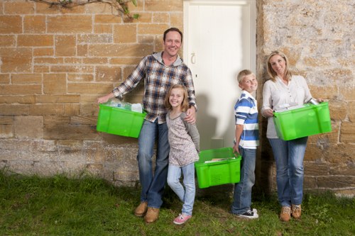 A family standing outside a stone and brick building on a grassy area, holding bright green plastic storage boxes. The father, dressed in a plaid shirt and jeans, is smiling at the camera while holding a box in his right hand. A young girl in a grey patterned dress and pink shoes stands beside him, also smiling, and holding a box. To the right, a boy in a blue and white striped shirt and jeans stands with a box in front of him, looking towards the camera. An older woman wearing a white blouse and jeans, with sandals, holds a similar green box and is positioned further right against the wall. The scene appears to be in a suburban or garden setting, with the family involved in rubbish or garden clearance, which aligns with services offered by Garden Clearance Surbiton.