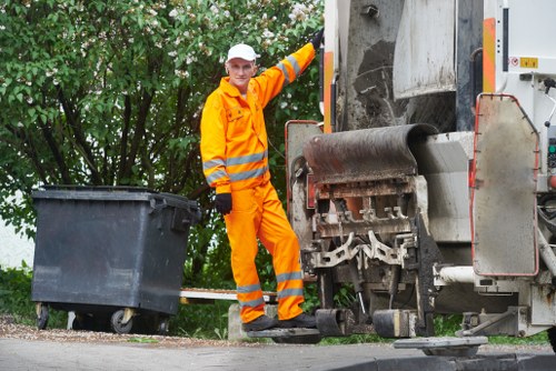 A waste collection worker wearing a high-visibility orange uniform, including jacket and trousers with reflective stripes, standing next to a large industrial rubbish compactor vehicle on a residential street. The worker, also wearing a white helmet and black gloves, is pointing towards the compactor's loading area, which has a black waste bin positioned nearby for collection. The background features green leafy trees and a paved pavement, suggesting a suburban environment typical of areas in Surbiton. The scene captures the process of rubbish collection, with the vehicle's robust metal surface and mechanical components clearly visible, emphasizing the practical aspect of waste management services provided by Garden Clearance Surbiton. Lighting indicates natural daylight, creating an evenly lit scene. This image reflects typical rubbish removal operations in the local area, highlighting the professional approach to waste disposal within the community.